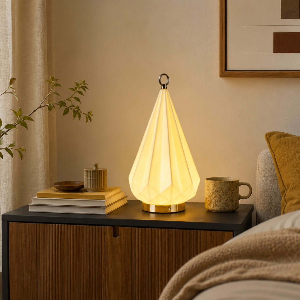 Clérachic cone lamp glowing on a fluted wood cabinet, with a stack of books, a speckled mug and a mustard cushion behind.