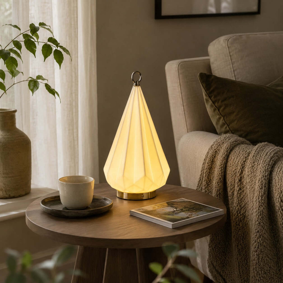 Faceted ceramic lamp from Clérachic on a round wooden side table, beside a cup and saucer and a magazine, next to a grey armchair.