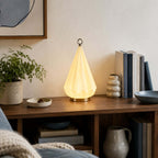 Lit Clérachic cone lamp on a wooden shelf unit with books, a small bowl and a stacked ceramic vase, with a plant at the left.