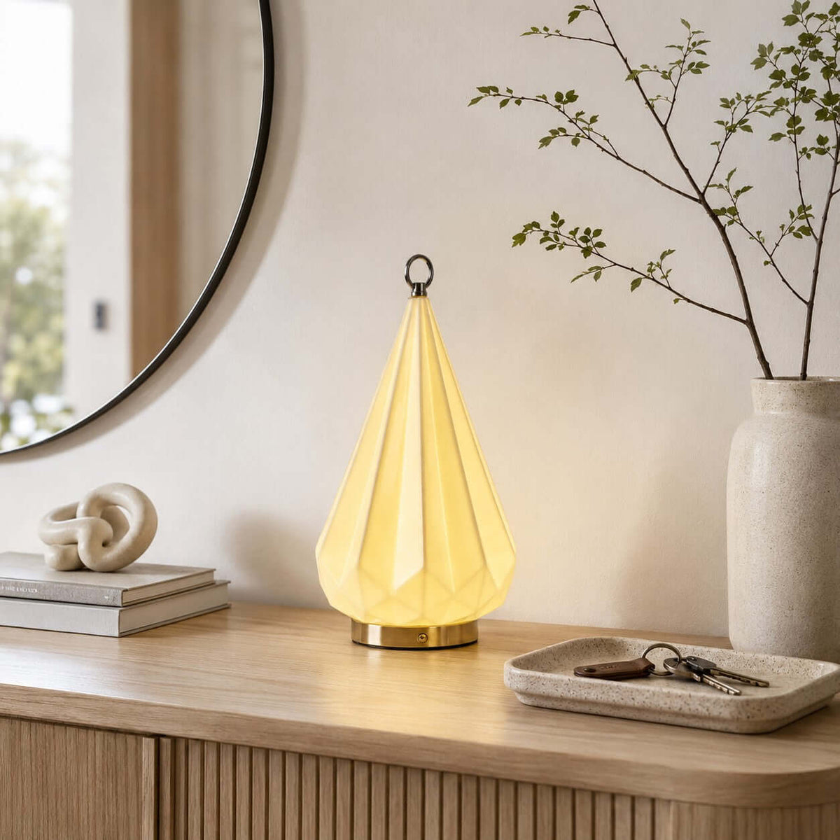 Clérachic faceted lamp lit on a fluted oak sideboard, with a key tray, a knotted sculpture, books and a tall vase with branches.