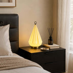 Faceted cone lamp from Clérachic glowing on a black bedside table beside a reed diffuser, a book and a small dish.