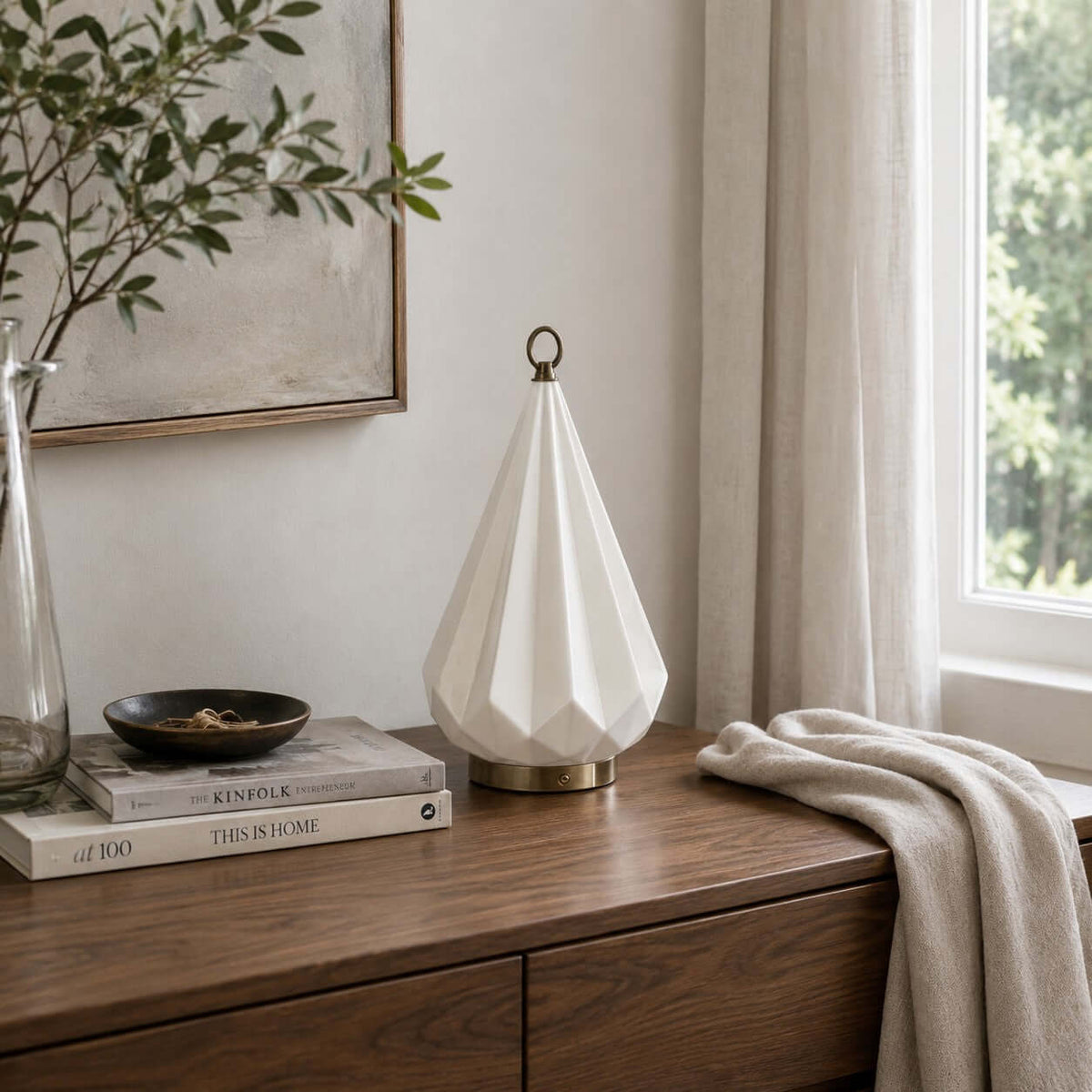 White faceted ceramic Clérachic table lamp, unlit, on a walnut sideboard by a window, with books, a dark bowl and a folded throw.