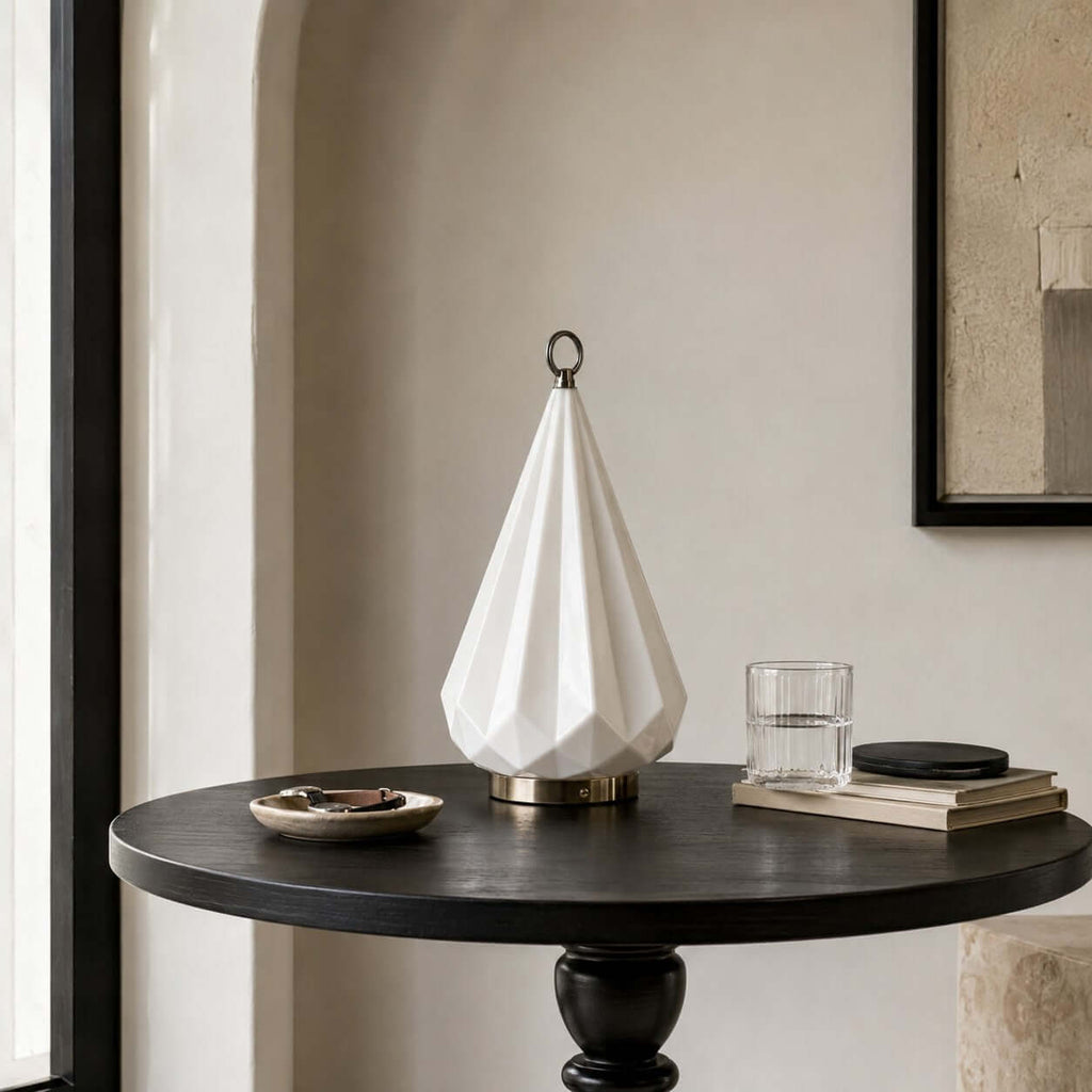 Unlit white faceted lamp from Clérachic on a round black side table, with a glass tumbler and a stack of books in a corner by an arch.
