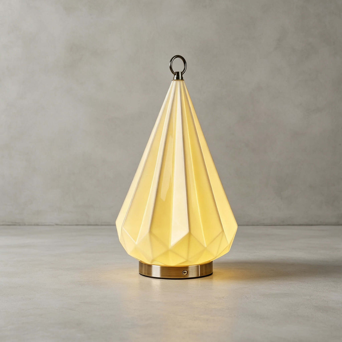 Clérachic faceted cone lamp lit warm yellow on a grey concrete-look floor, against a plain grey wall.