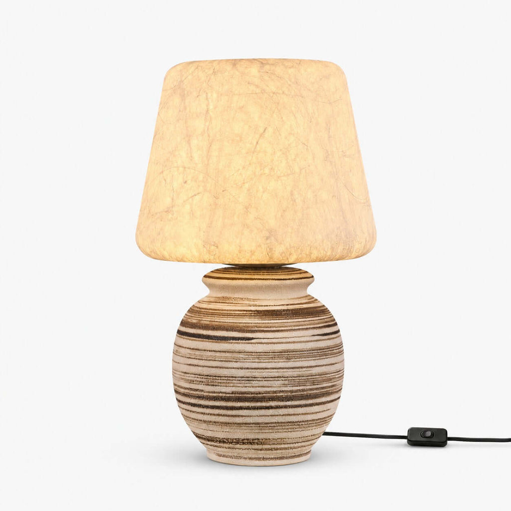 This Clérachic table lamp on a plain white backdrop, a tapered pale shade over a rounded ceramic base with brown and cream stripes and an inline cord switch.
