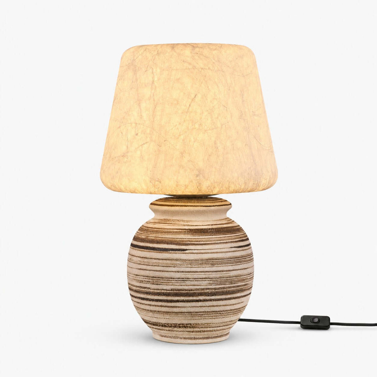 This Clérachic table lamp on a plain white backdrop, a tapered pale shade over a rounded ceramic base with brown and cream stripes and an inline cord switch.