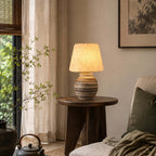 On a round dark wood side table next to a cream armchair, this Clérachic table lamp glows beside a sheer curtain and a leafy branch.