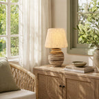 On a rattan-fronted cabinet by a green-framed window, this Clérachic table lamp stands next to a stack of books and a shallow bowl.