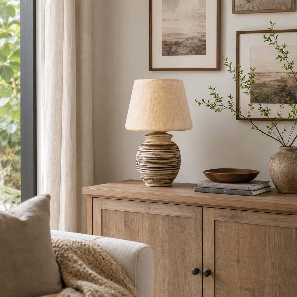 Unlit on a pale oak sideboard, this Clérachic table lamp shows its cream shade next to a wooden bowl and a stack of books.