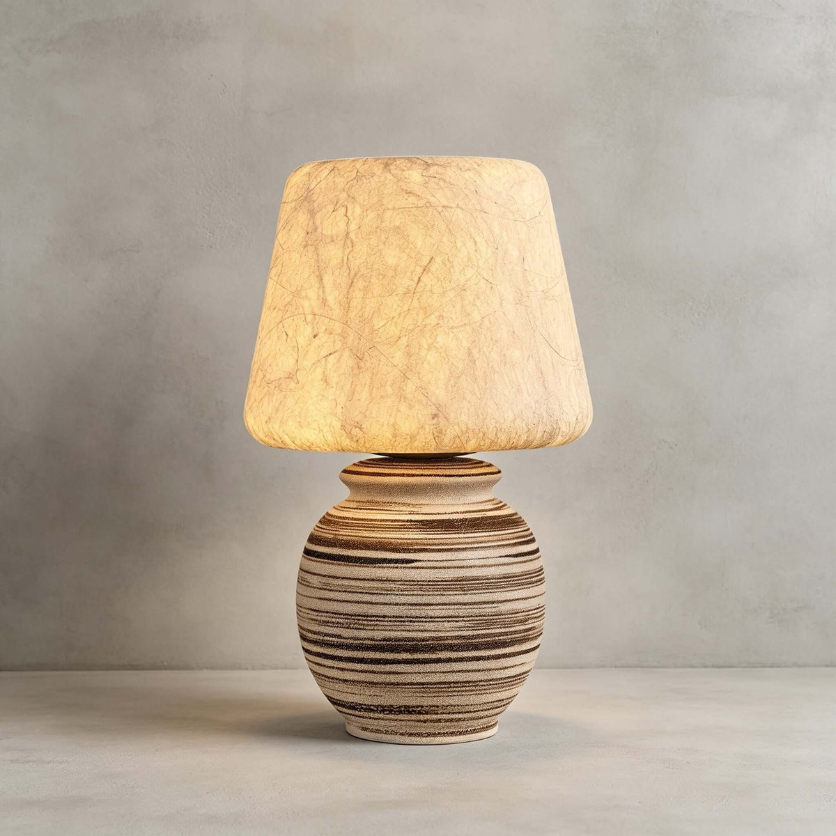 Close view of this Clérachic table lamp on a pale concrete-look backdrop, its fibrous shade glowing above the striped ceramic base.