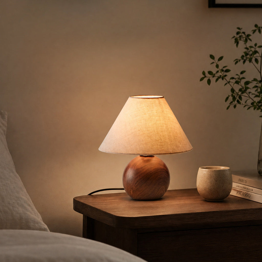 A lit Clérachic table lamp with a linen shade glowing warm on a dark wood bedside table, a small cup and books beside it.