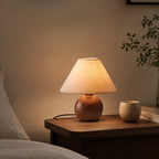 A lit Clérachic table lamp with a linen shade glowing warm on a dark wood bedside table, a small cup and books beside it.