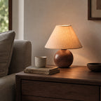 This Clérachic table lamp, its round walnut-grain base under a lit linen shade, on a nightstand next to a grey armchair.