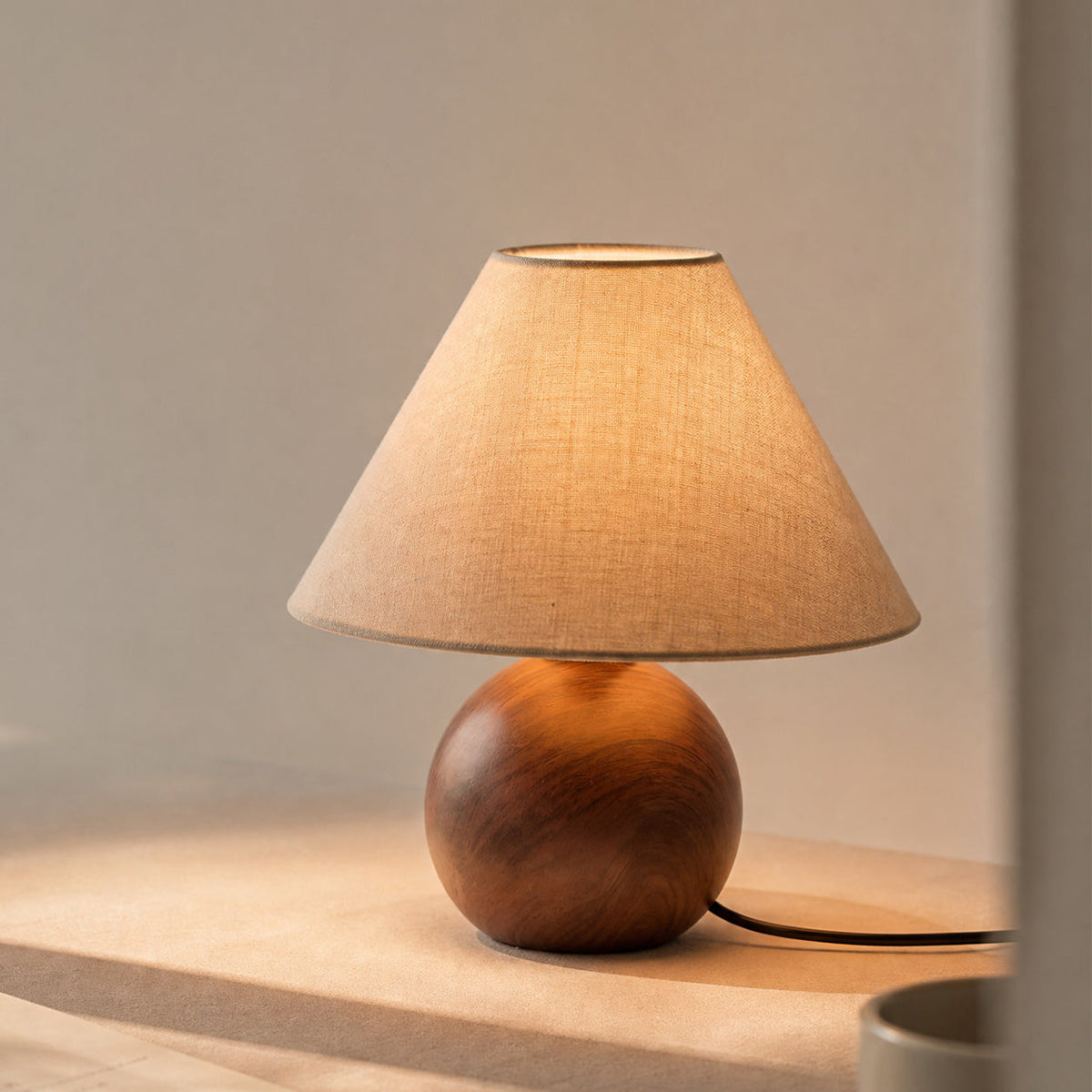 Clérachic linen-shaded table lamp lit on a pale surface in soft side light, its walnut wood-grain ball base in front of a grey wall.