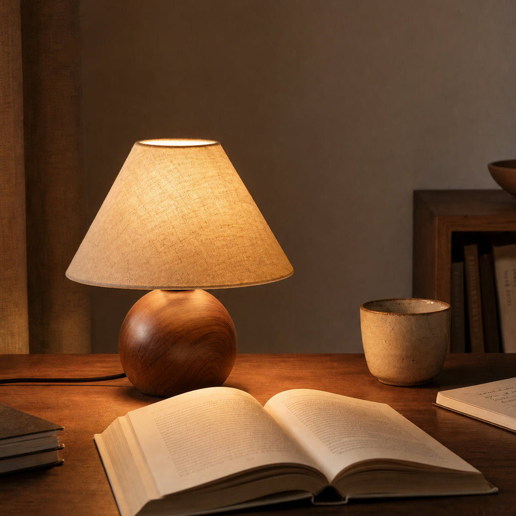 A Clérachic lamp glowing amber on a wooden desk, in front of an open book, a ceramic cup and a small bookshelf.
