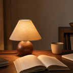 A Clérachic lamp glowing amber on a wooden desk, in front of an open book, a ceramic cup and a small bookshelf.