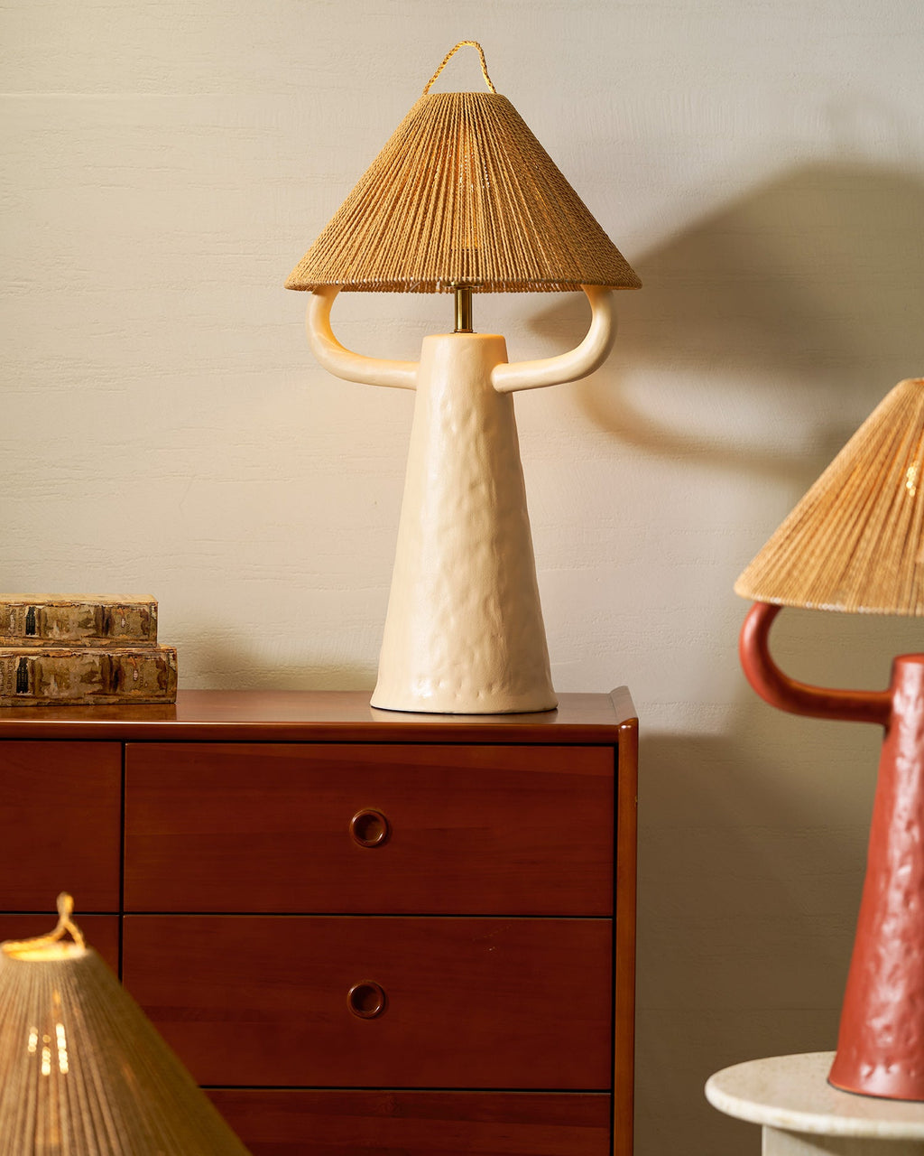 Cream ceramic Clérachic table lamp with curved arms and a rope shade on a walnut cabinet, with a stack of books to the left.