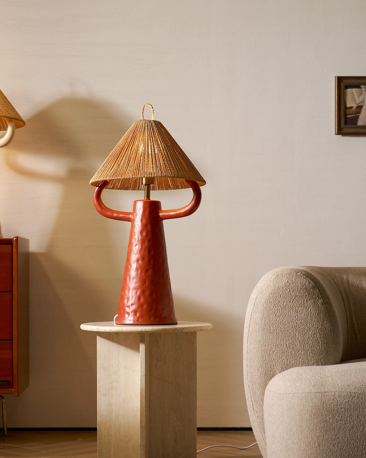 A terracotta ceramic lamp with a rope shade stands on a stone side table beside a cream boucle armchair; from Clérachic.