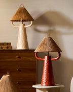 Terracotta table lamp with a woven shade at the front, a cream lamp on a walnut chest behind it, in a warm beige room; Clérachic.