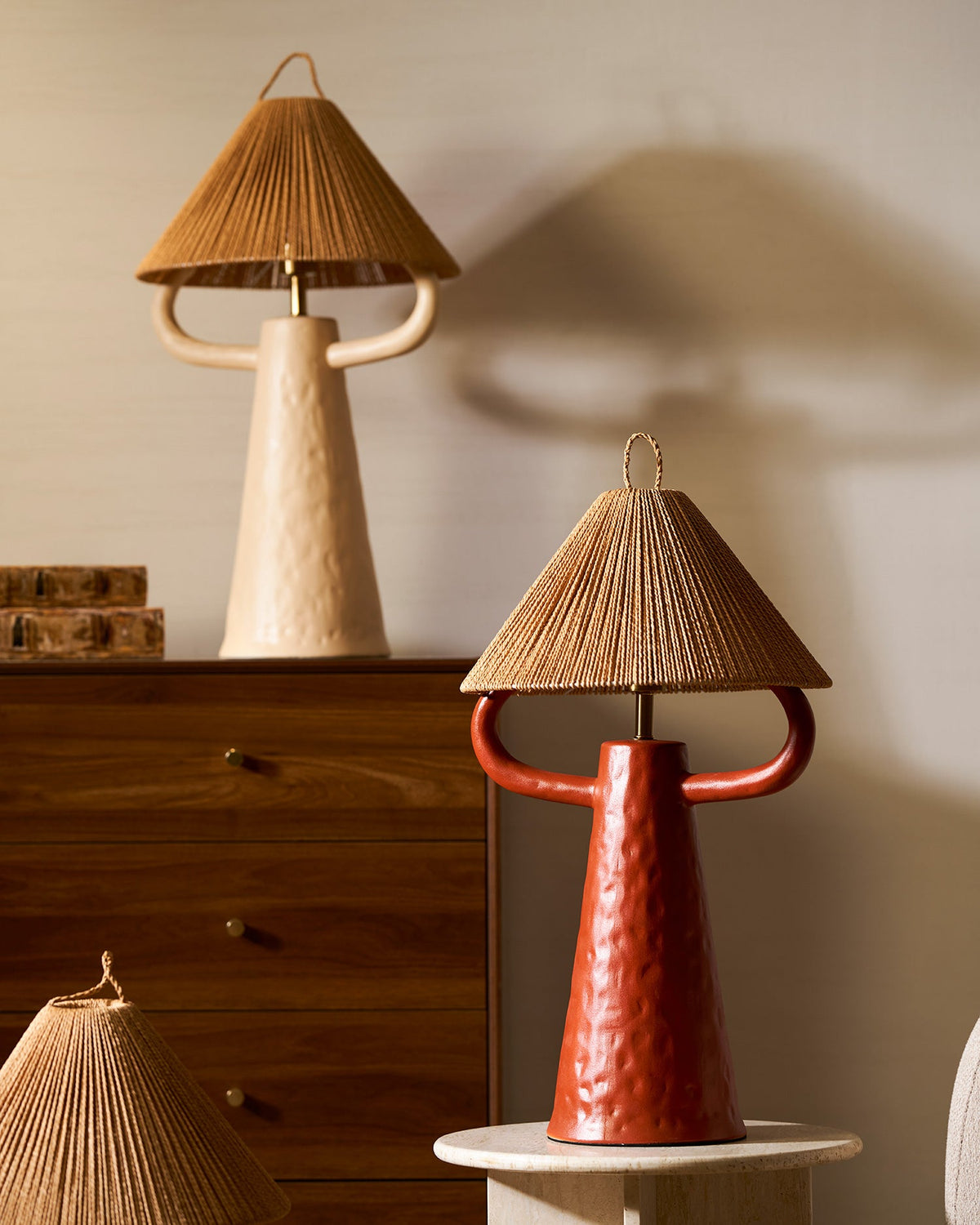 Terracotta table lamp with a woven shade at the front, a cream lamp on a walnut chest behind it, in a warm beige room; Clérachic.