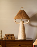 Close view of the cream Clérachic table lamp on a walnut cabinet, its dimpled ceramic body and two arms under a woven rope shade.