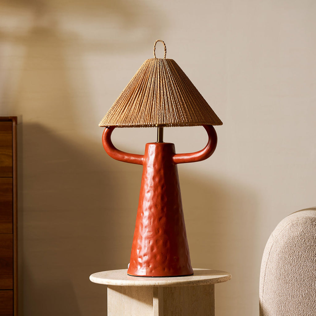 Terracotta ceramic lamp on a round stone table against a warm wall, with a rope shade and a boucle chair at the edge; from Clérachic.