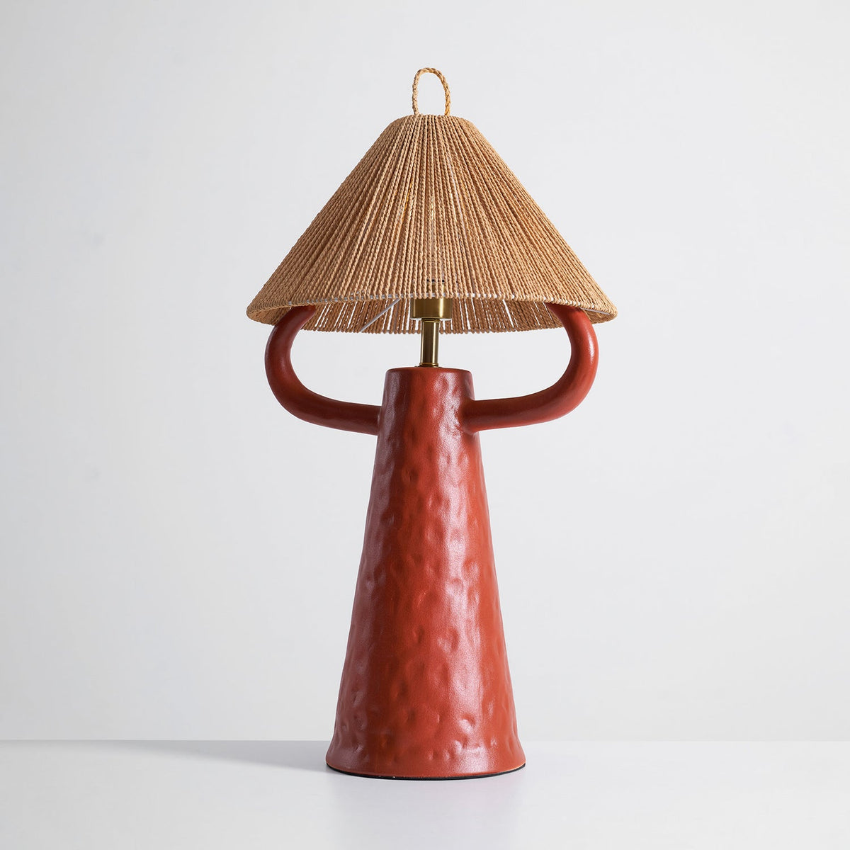 Terracotta Clérachic table lamp on a pale grey surface, the hammered-looking ceramic glazed deep red-orange below a woven rope shade.
