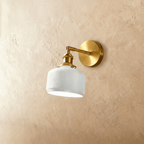 Clérachic wall lamp with a brass round backplate, angled arm and white ribbed ceramic shade, lit, on a warm plaster wall.