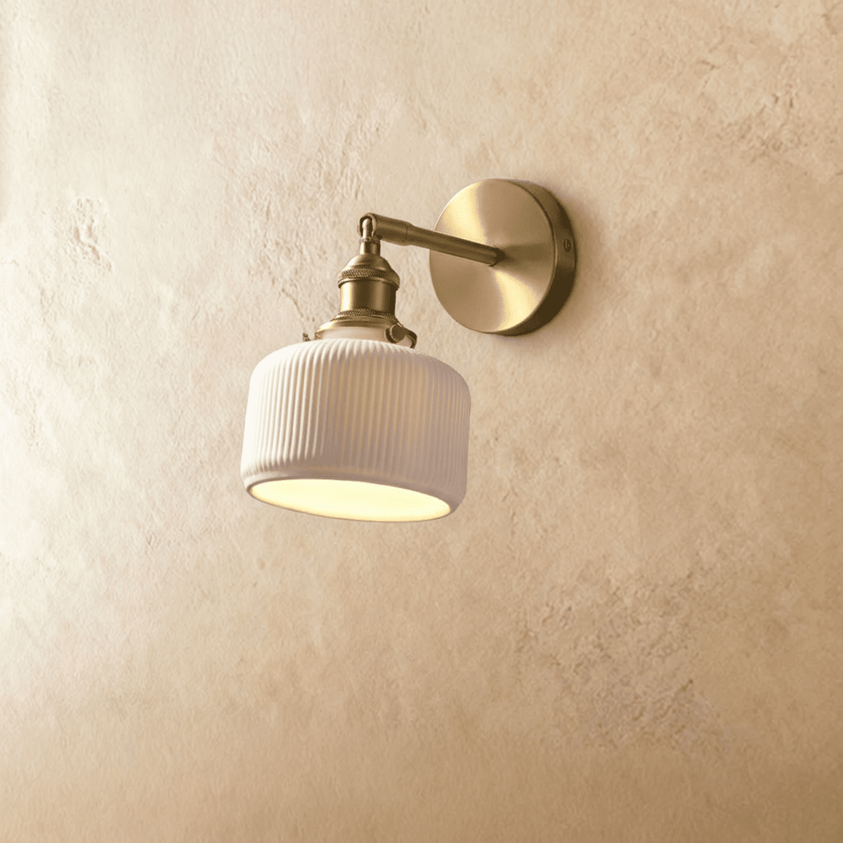 Clérachic wall lamp in brushed brass with a white ribbed ceramic shade, softly lit from below, against textured sand-coloured plaster.