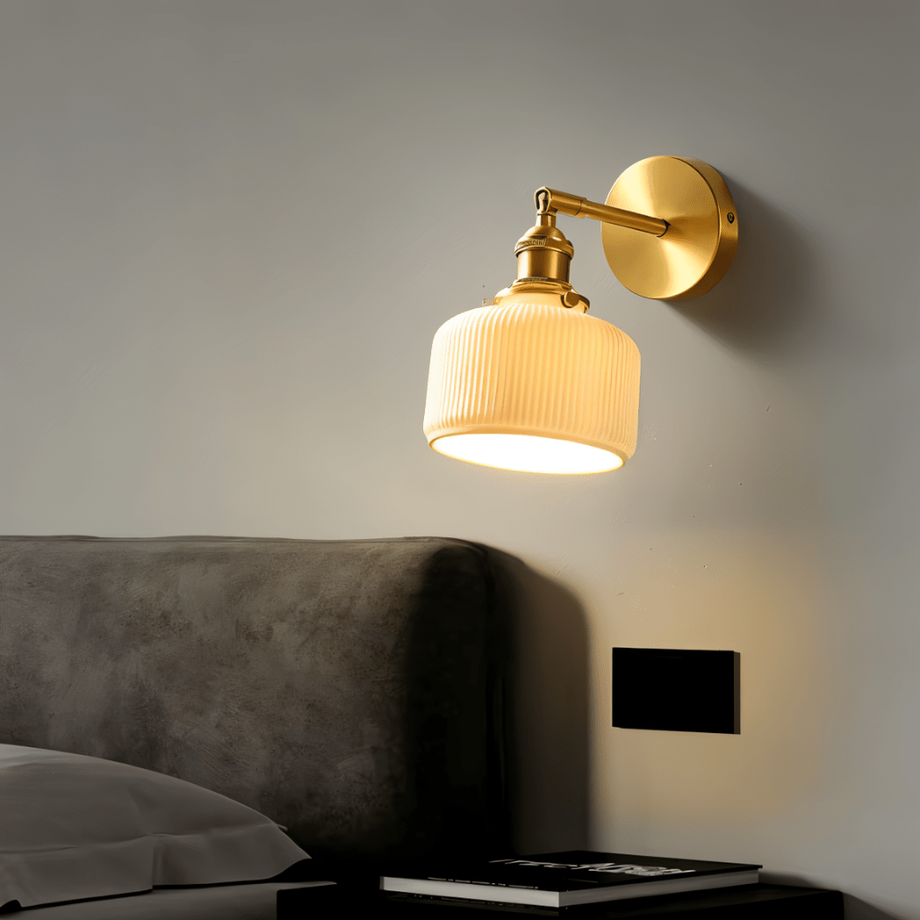 Clérachic brass wall lamp with a ribbed ceramic shade lit above a grey upholstered headboard and a black bedside table.