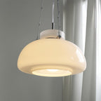 Close view of a lit Clérachic white glass dome pendant, with its stepped diffuser, chain links and chrome fittings, by a sheer curtain.