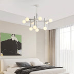 A Clérachic chandelier glowing high in a bedroom with a low white bed, grey bedding and a green portrait print on the wall.