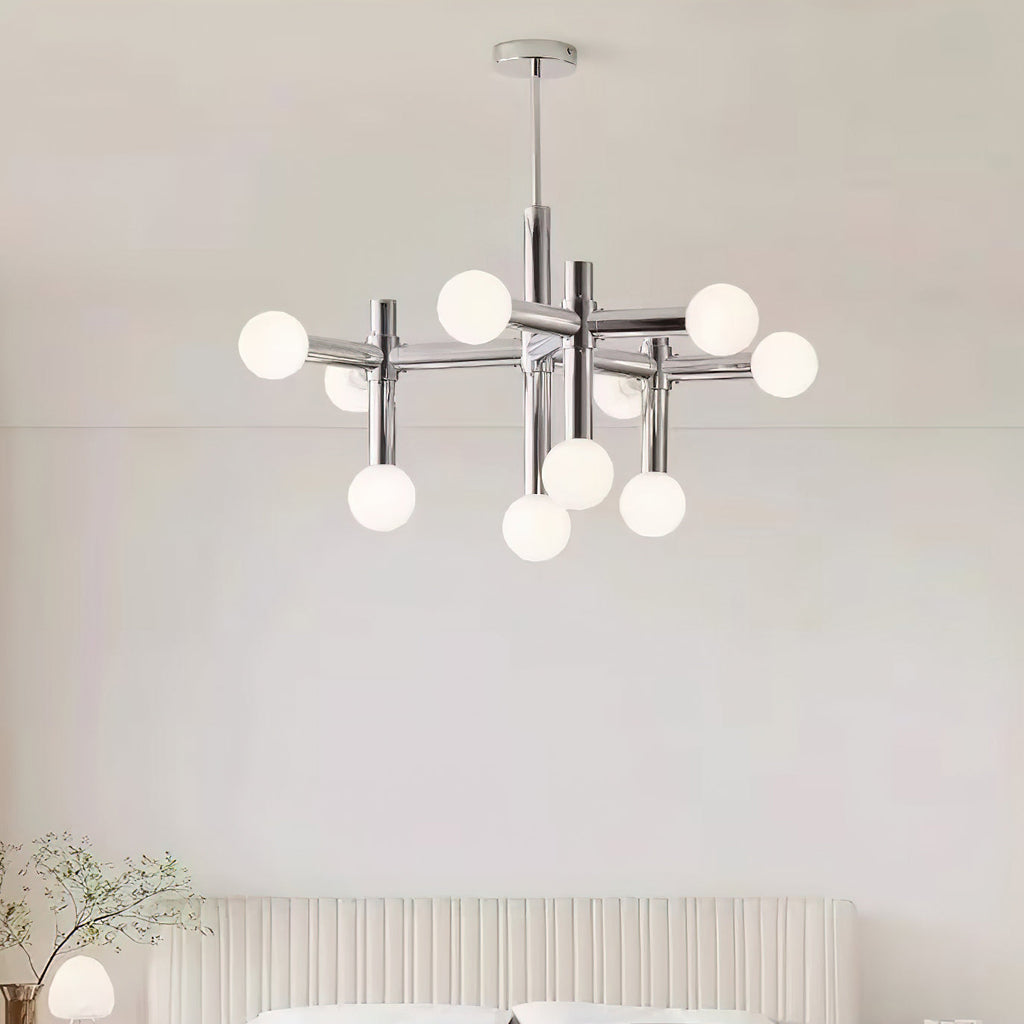 A Clérachic chandelier hanging above a bed with a fluted cream headboard, its chrome arms and white globes lit softly.