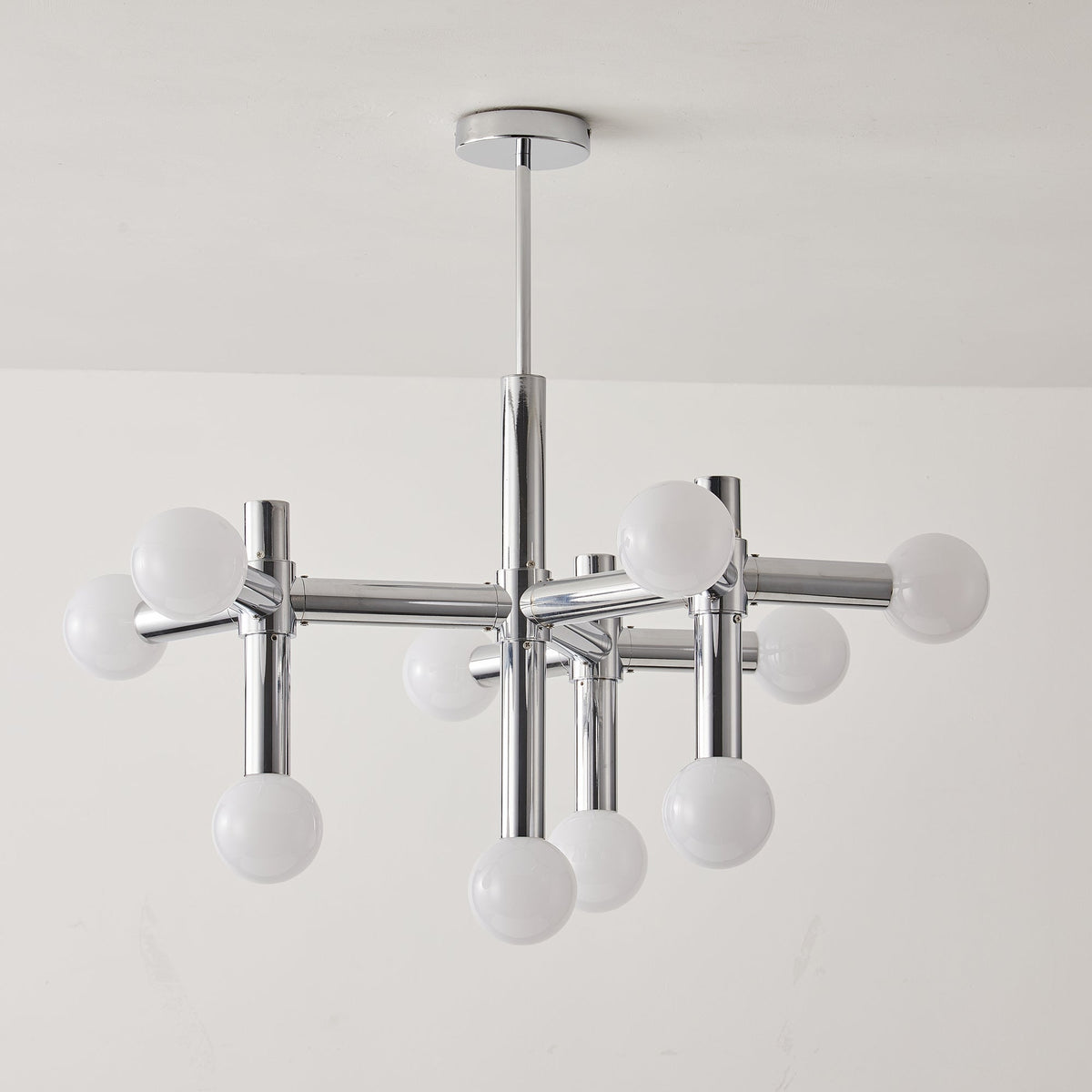 Unlit Clérachic chandelier against a pale wall, its chrome crossing arms and ten white globes hanging from a thin rod.