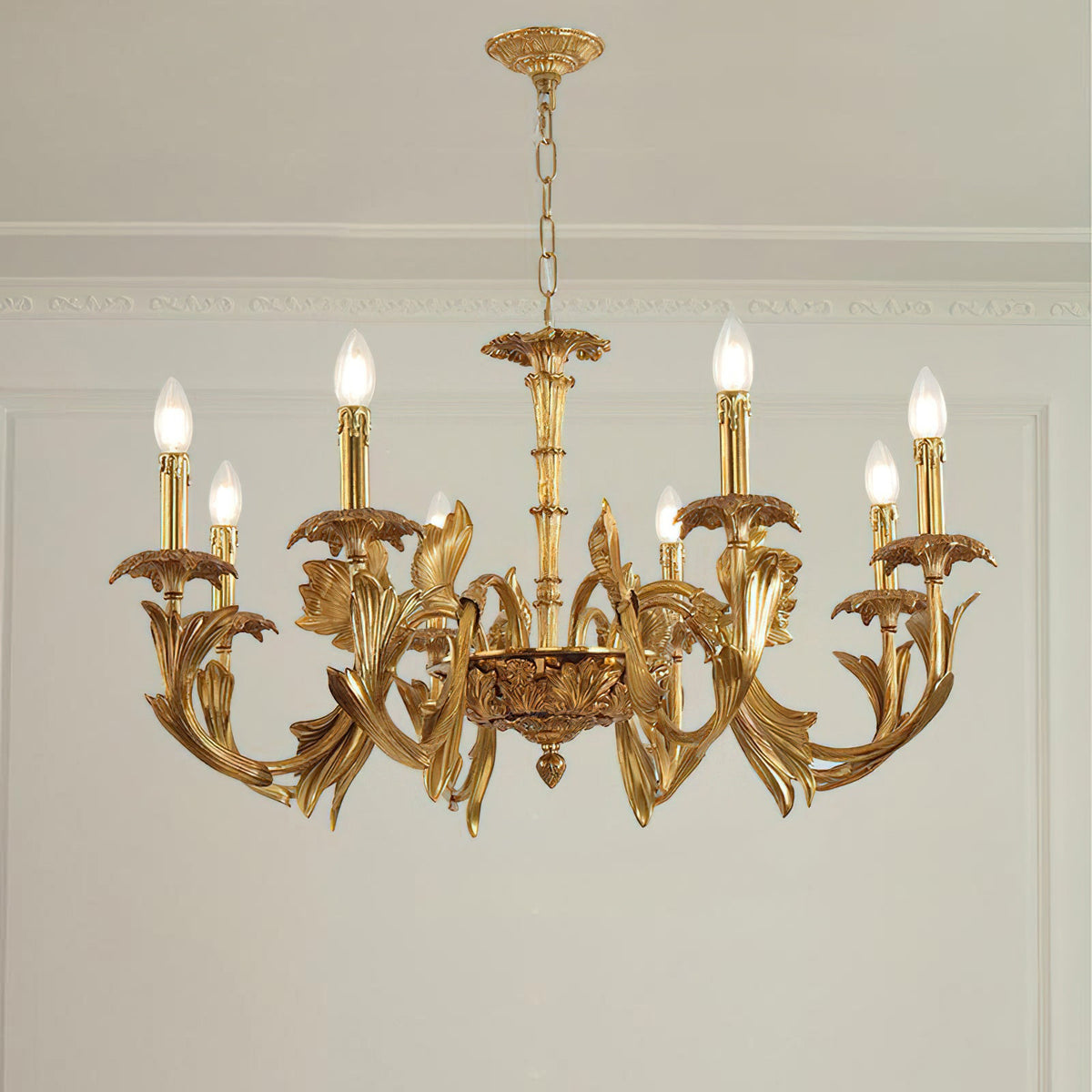Clérachic brass chandelier hanging in a room with a white panelled wall and cornice, its candle bulbs lit.