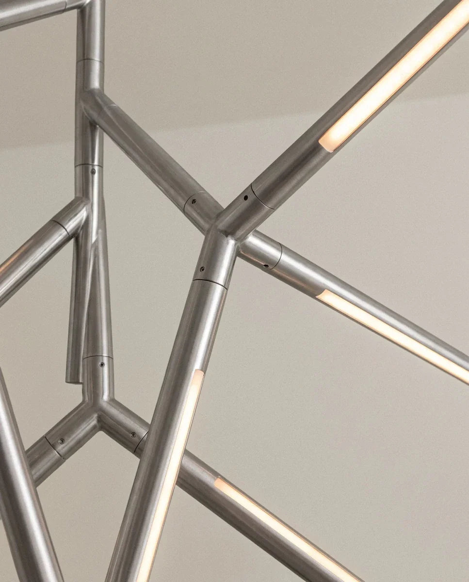 Clérachic close detail of chrome chandelier arms joined at angles, each rod carrying a glowing strip of warm light.