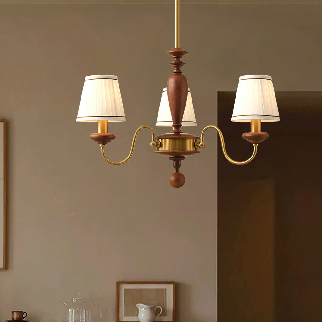 Three-light Clérachic chandelier in gold and walnut, its pleated shades lit against a taupe wall with framed pictures behind.