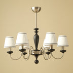 Six-light Clérachic chandelier in chrome and black, with white pleated shades trimmed in dark piping, on a warm beige backdrop.