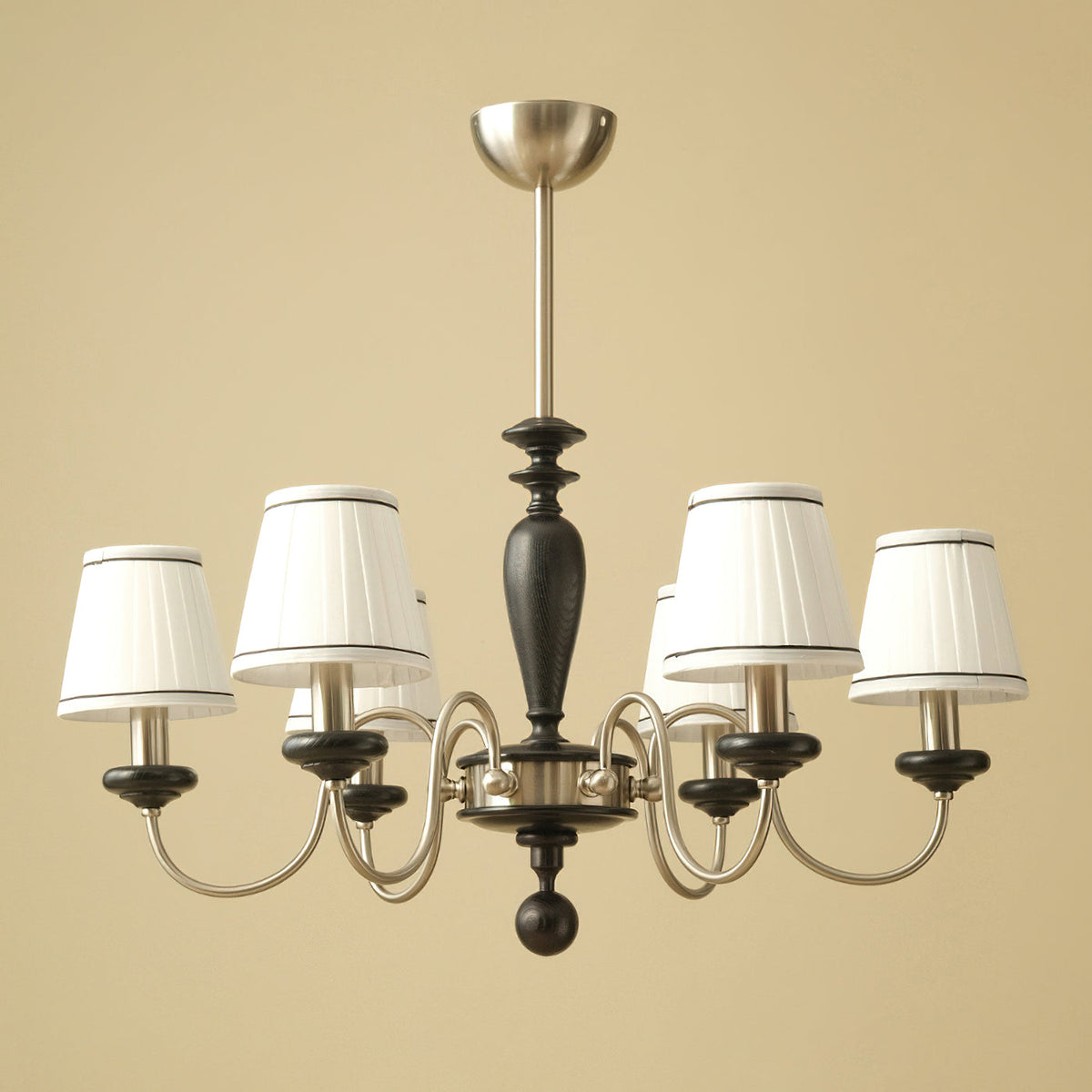 Six-light Clérachic chandelier in chrome and black, with white pleated shades trimmed in dark piping, on a warm beige backdrop.
