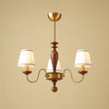 Three-light gold and walnut Clérachic chandelier, its S-shaped brass arms carrying lit pleated shades on a warm beige background.