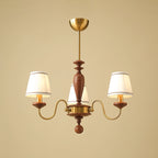 Three-light gold and walnut Clérachic chandelier, its S-shaped brass arms carrying lit pleated shades on a warm beige background.
