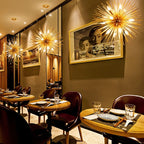 Several Clérachic gold spike chandeliers lit along a restaurant wall above round tables and leather chairs.