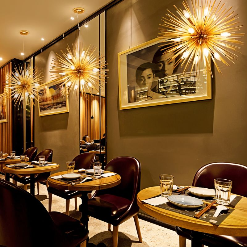 Several Clérachic gold spike chandeliers lit along a restaurant wall above round tables and leather chairs.