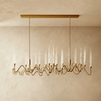 Clérachic linear brass chandelier on a rectangular canopy, with two dozen clear glass shades on wavy arms, against a plaster wall.