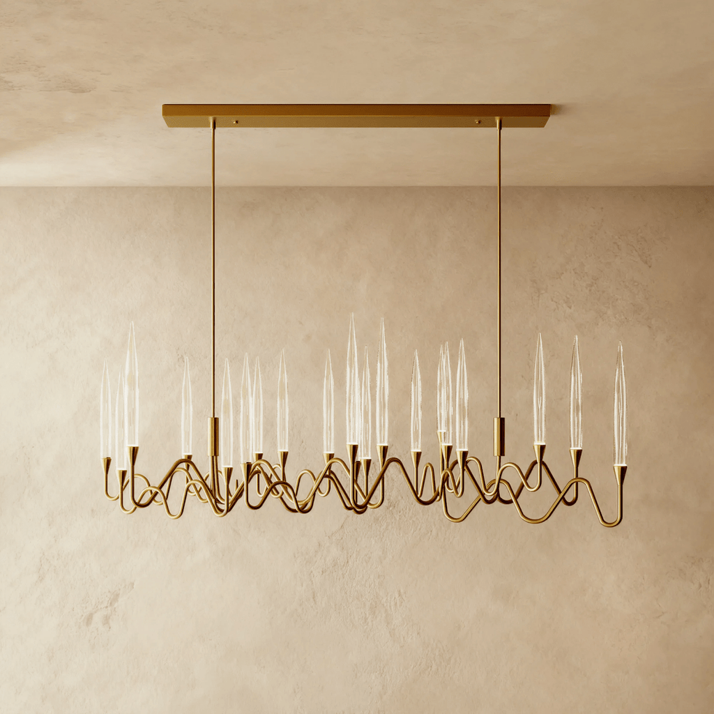 Clérachic linear brass chandelier with clear glass candle shades on rippling arms, hung from a rectangular canopy above a plaster wall.
