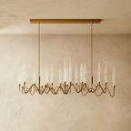 Clérachic linear brass chandelier with clear glass candle shades on rippling arms, hung from a rectangular canopy above a plaster wall.