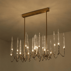 Clérachic linear brass chandelier lit warm on a rectangular canopy, its clear glass shades glowing against a softly lit ceiling.