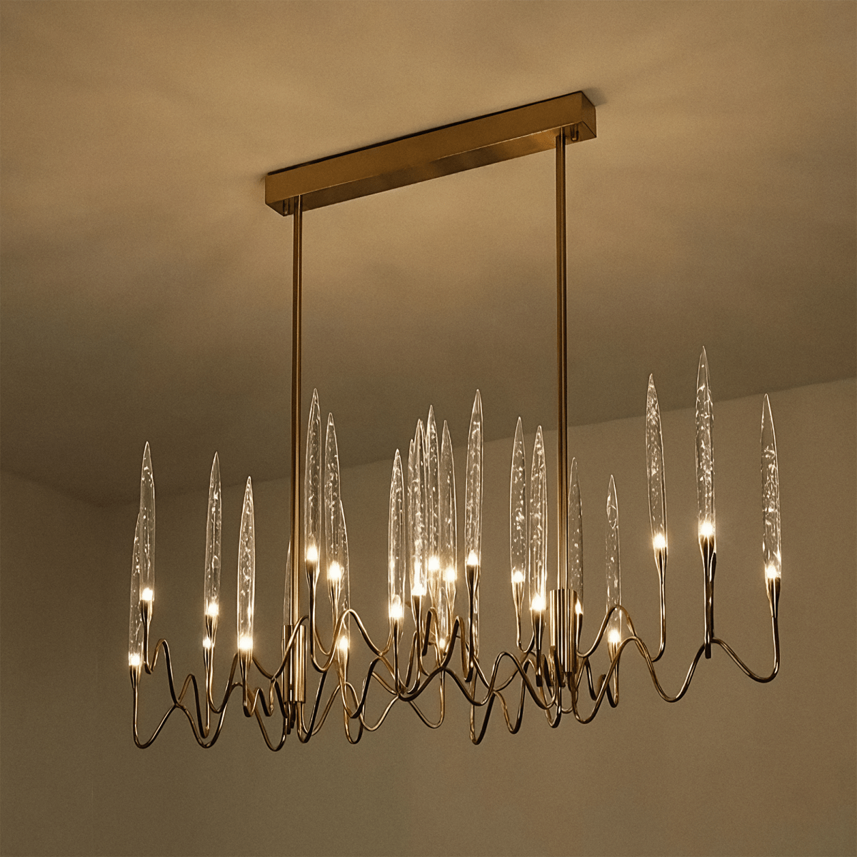 Clérachic linear brass chandelier lit warm on a rectangular canopy, its clear glass shades glowing against a softly lit ceiling.