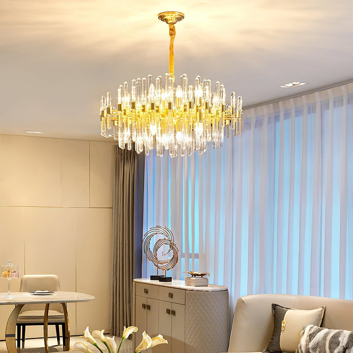 Warm light from this Clérachic brass chandelier fills a living and dining area, with sheer blue-tinted curtains and cream wall panels behind.