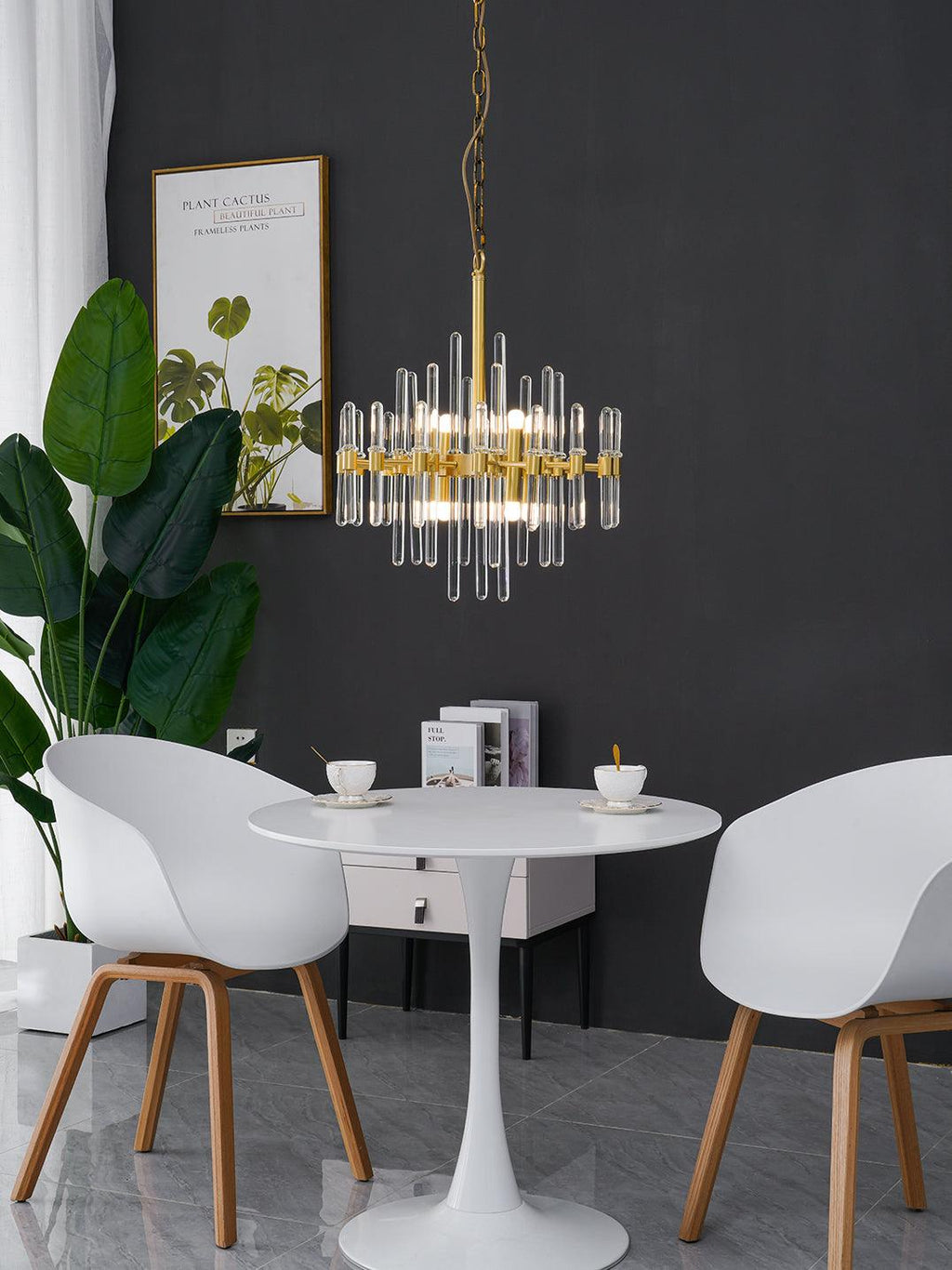 A compact Clérachic brass chandelier with clear glass rods hangs on a fine chain between a tall plant and white chairs.